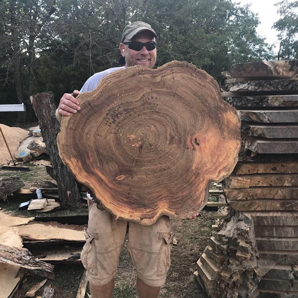 man holding slab in front of piles of cut wood